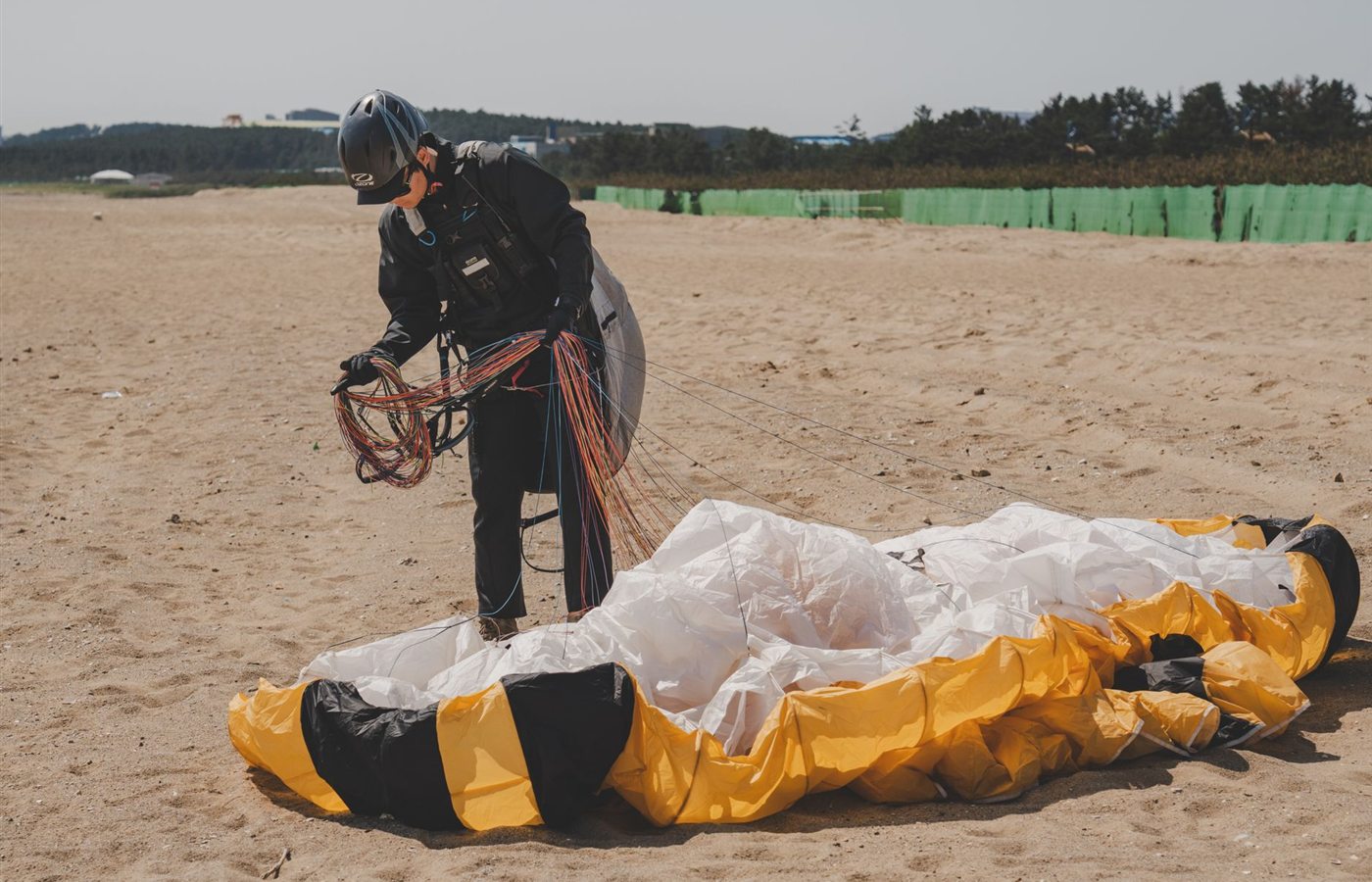 Individual packing a paraglider on a sunny beach, organizing lines and equipment.