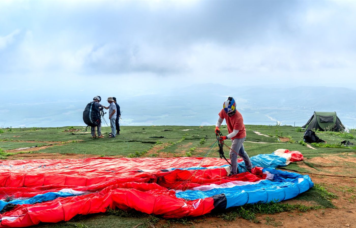 Paragliders setting up gear on a scenic plateau under a cloudy sky.